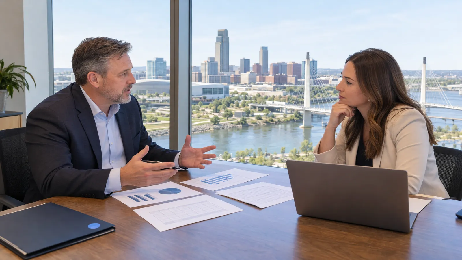 A business owner and a broker sit across a desk by a tall window with the Omaha riverfront and a pedestrian bridge in the distance, financial statements between them